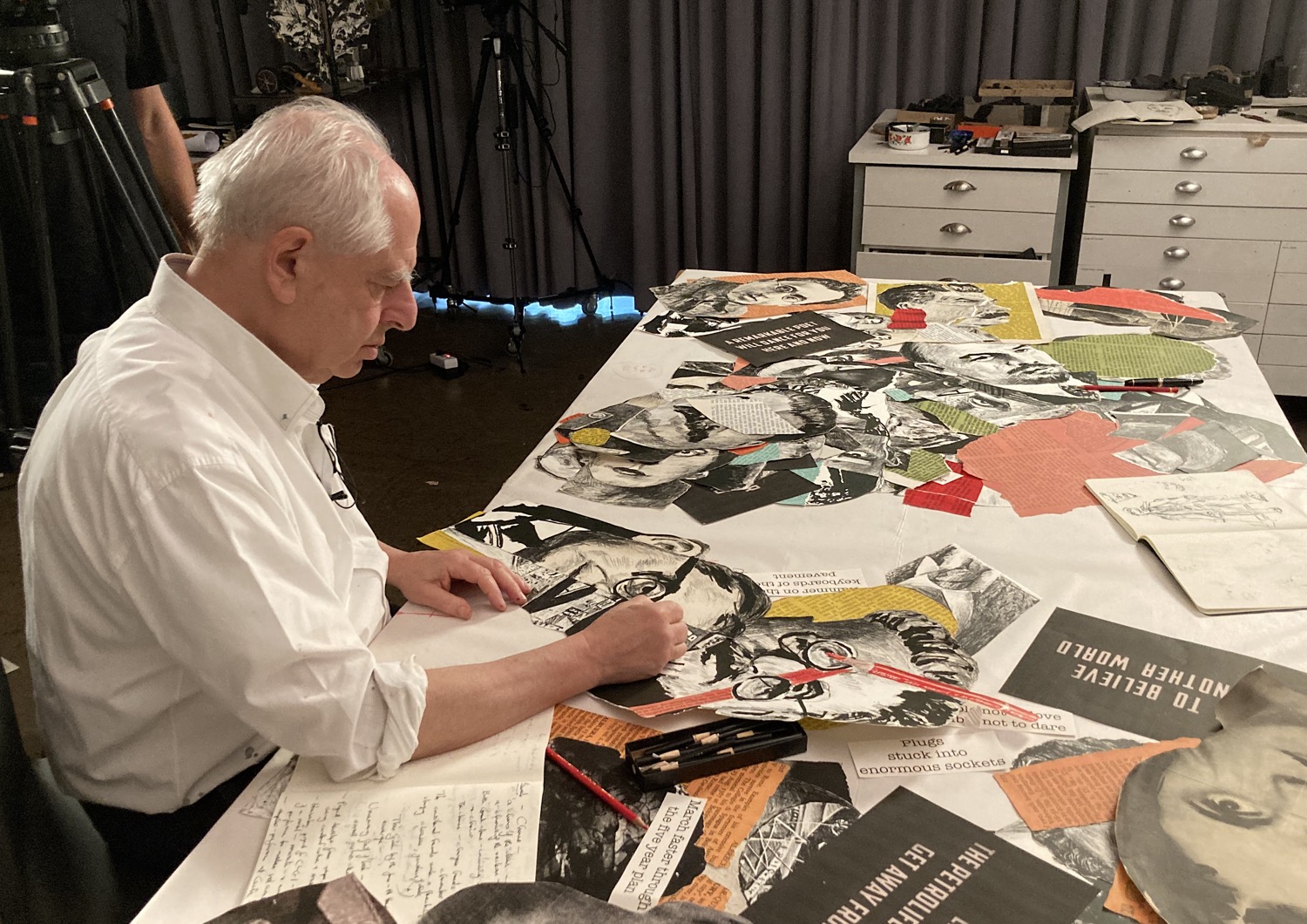 William Kentridge is seated in his studio with multiple collaged portraits in multi colours on the table